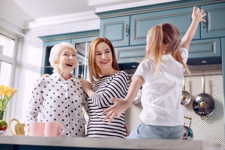 Interesting story. Adorable little girl sitting on the kitchen counter and sharing an exciting story with her beloved mother and grandmother while gesticulating a lotの写真素材
