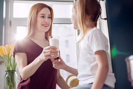 Drink it up. Beautiful young woman giving a glass of milk to her cute little daughter and smiling at her while the girl taking it with a smileの写真素材