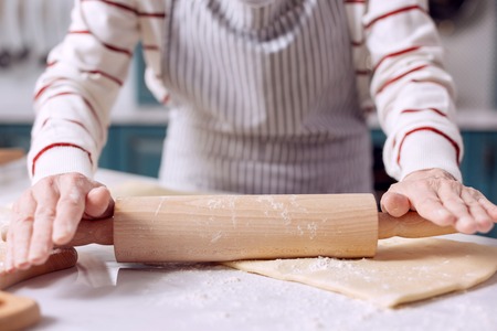 Future pie. The close up of delicate hands of an elderly lady in an apron using a rolling pin to roll out dough while making a pieの写真素材