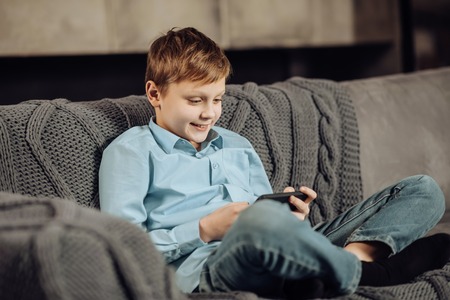 Calm pastime. Upbeat pre-teen boy sitting cross-legged on the sofa and playing on the phone while smiling pleasantlyの写真素材