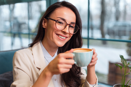 Charming aroma. Jolly cute pretty woman posing on the blurred background while looking at camera and drinking coffeeの写真素材