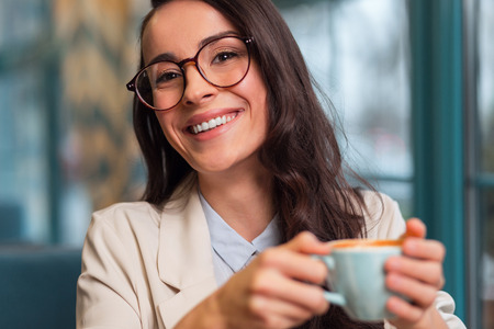 Nice day. Merry positive beautiful woman posing on the blurred background while grinning and carrying cup of coffeeの写真素材