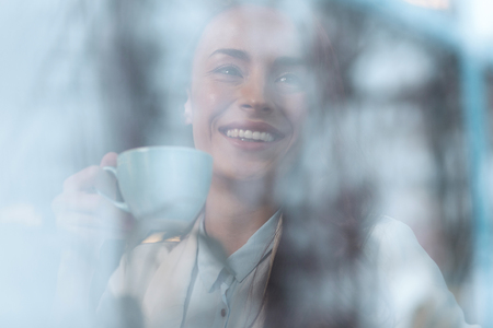 Warm cup. Pleasant satisfied glad woman enjoying coffee while smiling and holding cupの写真素材