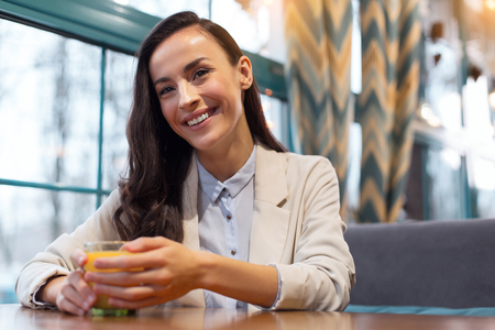 Essential vitamins. Attractive glad cute woman sitting at table while smiling and touching glass with juiceの写真素材