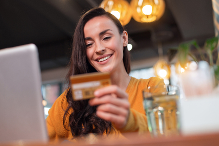 Payment everywhere. Low angle of sincere pleasant satisfied woman looking at credit card while smiling and posing on the blurred backgroundの写真素材