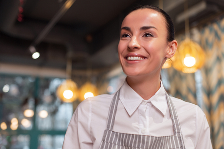 First customer. Portrait of dreamful jolly nice waitress posing on blurred background and smiling while looking asideの写真素材