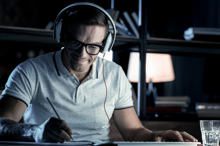Positive mood. Joyful millennial guy wearing headphones sitting at a table and grinning broadly while writing something down.の写真素材