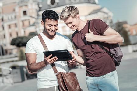 Look at this. Positive joyful young man standing together with his friend and talking while showing him an interesting pictureの写真素材