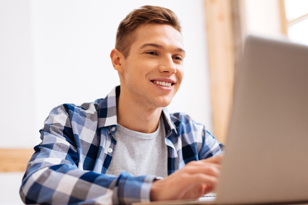 I feel great. Attractive content fair-haired boy working on his laptop and smiling and wearing a tartan shirtの写真素材