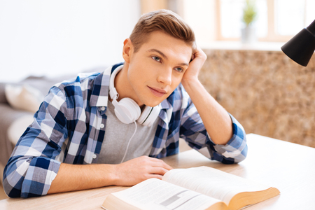 Thinking. Attractive inspired fair-haired boy smiling and sitting at the table with a book and thinkingの写真素材
