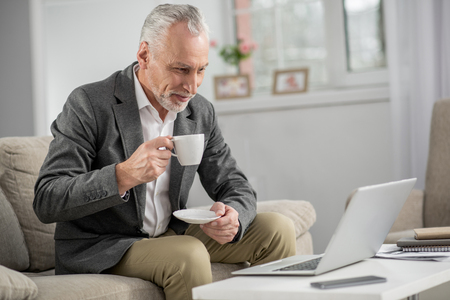 Keep your attention. Attractive businessman holding cup in right hand and keeping smile on his face while looking at computerの写真素材