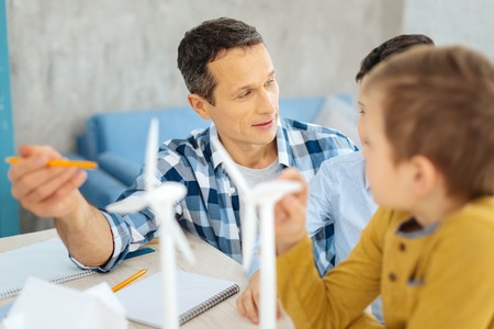 Adult discussion. Charming young man sitting at the table with his sons and discussing with them the advantages of wind turbines while pointing at themの写真素材