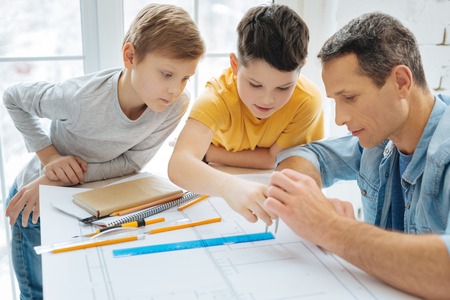 Easy lesson. Curious pre-teen boys leaning over their fathers work desk and learning how to use the compass while their father teaching them to do soの写真素材