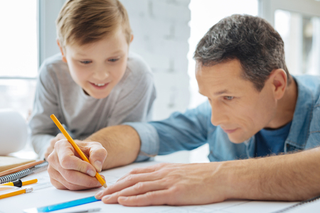 Fascinated by work. Pleasant curious pre-teen boy sitting at the table next to his father and watching him draw a blueprint with the pencil and rulerの写真素材