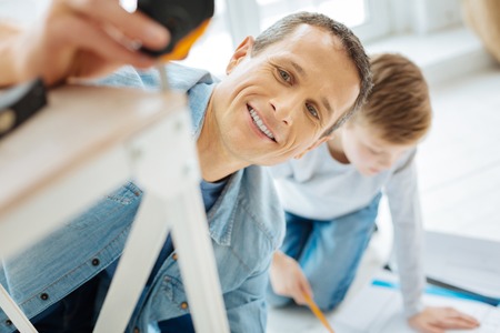 Happy carpenter. The close up of a cheerful young man measuring the leg of a table with the help of a measuring tape and smiling while his son studying the blueprintの写真素材
