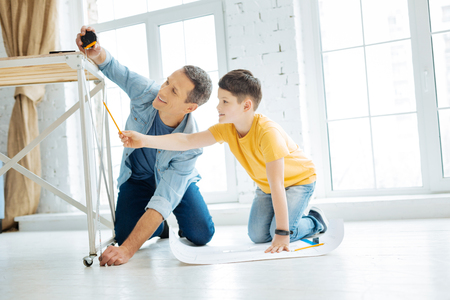 Best team. Cheerful charming young father working on a table construction together while the man measuring the length of a table leg and the boy checking its correctnessの写真素材