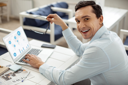Good job. Handsome joyful dark-haired man smiling and working on his laptop and holding his glasses while sitting at the tableの写真素材