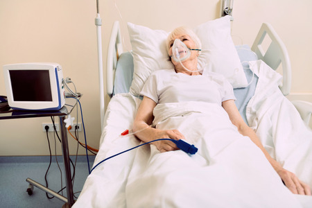 Healing process. Poor retired woman lying in a hospital bed with a drop counter and a heart rate medical equipment on her hand.の写真素材