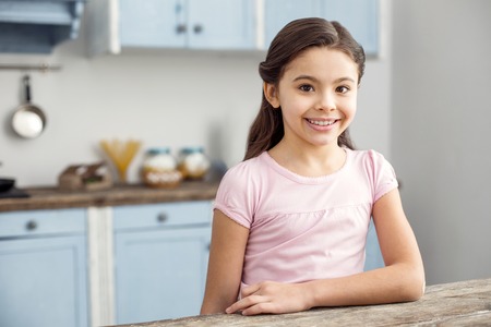 Happiness inside me. Pretty cheerful dark-haired little girl smiling and sitting at the table in the kitchen and wearing a pink shirtの写真素材