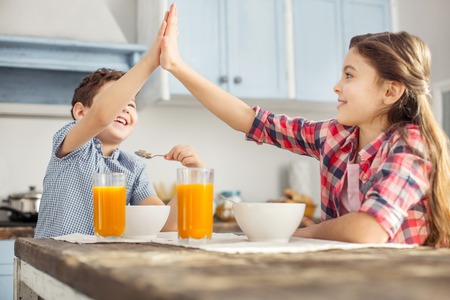 Good job. Beautiful inspired little dark-haired girl smiling and looking at her brother while they having breakfast and giving him a high-fiveの写真素材