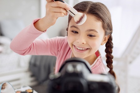 Favorite hobby. The close up of a pretty pre-teen girl grinning at the camera and applying powder to her forehead while filming a video tutorialの写真素材