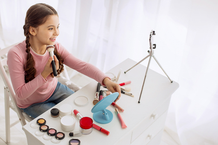 Wannabe makeup artist. Charming upbeat pre-teen girl posing for the camera and reviewing makeup products live, broadcasting it in the Internetの写真素材