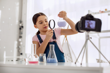 Talented chemist. Lovely pre-teen girl looking at a flask with a chemical in it through a magnifying glass while recording herself for a video blogの写真素材