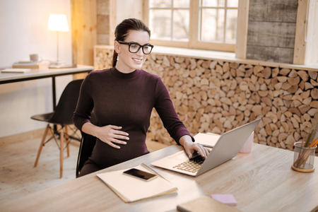 Business documentation. Charming smart pregnant businesswoman wearing glasses and typing on laptop while embracing bellyの写真素材