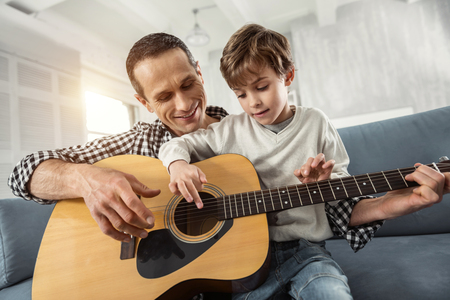 Happiness. Attractive concentrated fair-haired boy learning to play the guitar while sitting on the couch and his father smilingの写真素材