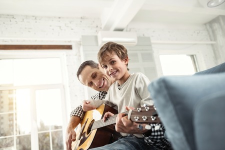 Good mood. Handsome smiling fair-haired boy learning to play the guitar while sitting on the couch and his father smilingの写真素材