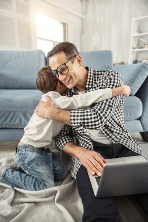 I cherish you. Handsome exuberant dark-haired father sitting on the floor and working on his laptop and his son hugging himの写真素材