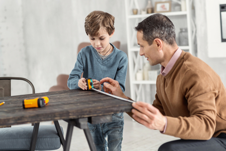 Measuring. Good-looking concentrated little fair-haired boy holding a measuring tape and measuring the table and his daddy helping helpの写真素材