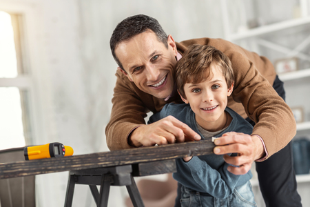 Caring father. Attractive exuberant little fair-haired boy holding a measuring tape and measuring the table and his daddy helping helpの写真素材