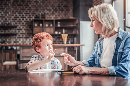 Time to study. Nice smart aged woman sitting at the table and talking to her grandson while teaching himの写真素材