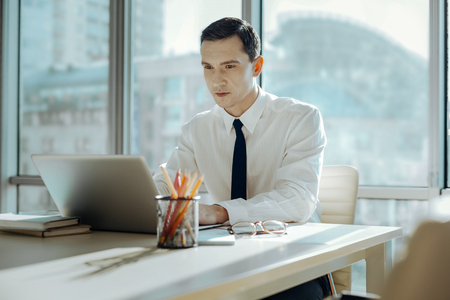 Immersed in work. Handsome black-haired young man sitting at the table in his office and working on his laptop, being completely immersed in workの写真素材