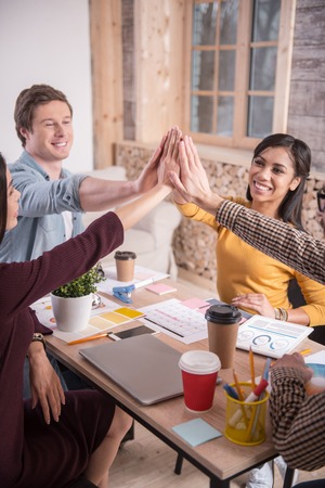 This is success. Nice positive delighted people sitting around the table and giving high five while...