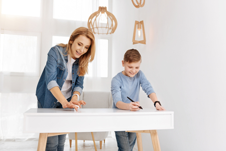 Pleasant entertainment. Delighted positive happy mother and son standing near the table and using modern technology while having fun togetherの写真素材