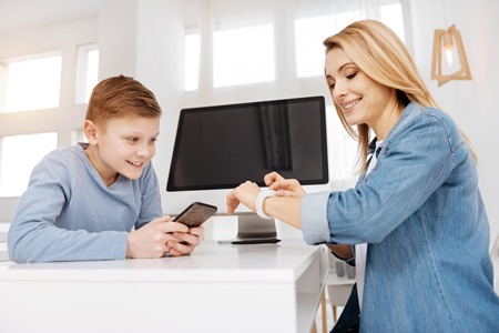 Modern generation. Happy nice young woman smiling and holding his smartphone while looking at his mothers smartwatchの写真素材