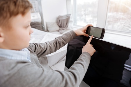 Innovative smartphone. Pleasant nice young boy holding his smartphone and looking at its screen while using modern technologyの写真素材