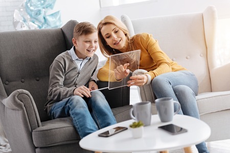 Pleasant interaction. Delighted nice positive woman sitting on the sofa and talking to her son while showing him something interesting on the tabletの写真素材