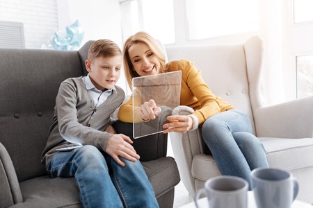 Pleasurable time. Joyful positive mother and son sitting on the sofa and looking at the tablet screen while resting at homeの写真素材