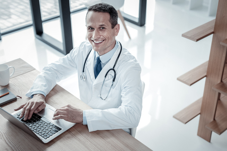 Everything will be fine. Joyful pleasant skilled medic sitting in the cabinet smiling and using the laptop.の写真素材
