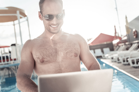 Positive attitude to work. Extremely happy millennial guy wearing sunglasses grinning broadly while working near a swimming pool and sunbathing at the same time.の写真素材