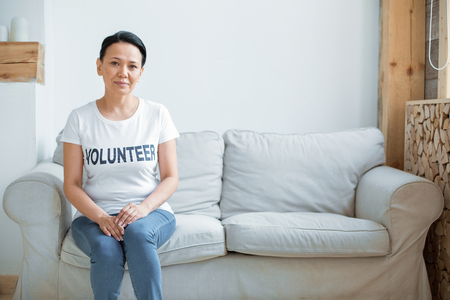 Time to work. Asian attractive female volunteer looking at camera and sitting on sofa while putting hands on kneesの写真素材