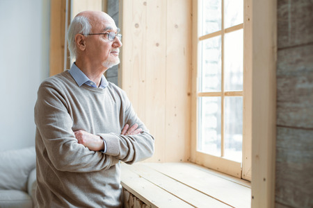 Pensive mood. Thoughtful attractive senior man gazing through window while crossing hands and...
