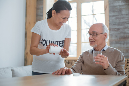 Regular cold. Beautiful positive volunteer standing while proposing pills to senior man who holding glass go waterの写真素材