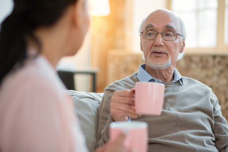 Herbal tea. Vigorous enthusiastic senior man talking with caregiver while drinking tea and rising mugの写真素材