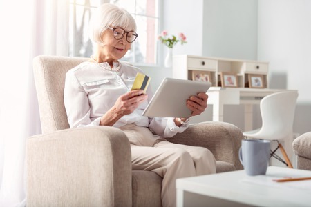 Easy and convenient. Adorable elderly woman sitting in the comfortable armchair and doing online shopping, ready to pay with her credit cardの写真素材