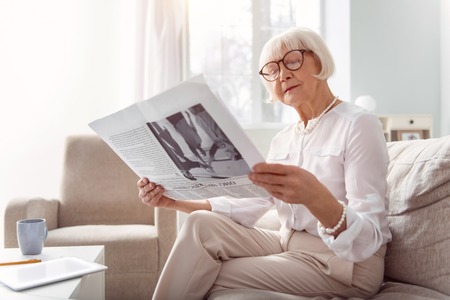 Interesting article. Beautiful senior lady sitting on the sofa in the living room and reading a newspaper, paying attention to itの写真素材
