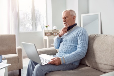 Enjoy reading. Handsome elderly man sitting on the couch and reading from the laptop screen while...